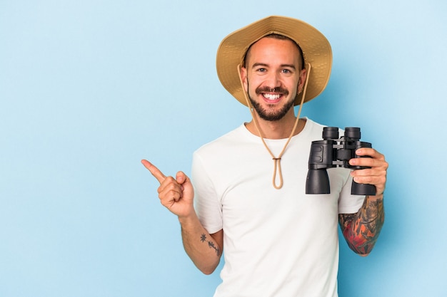 Photo jeune homme de race blanche avec des tatouages tenant des jumelles isolées sur fond bleu souriant et pointant de côté, montrant quelque chose dans un espace vide.