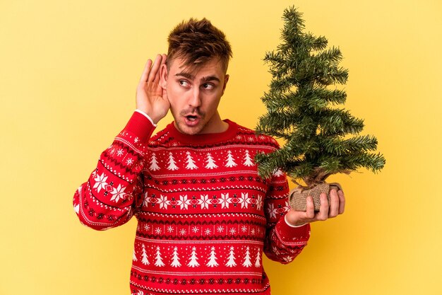 Jeune homme de race blanche tenant un arbre de Noël isolé sur fond jaune essayant d'écouter un potin.