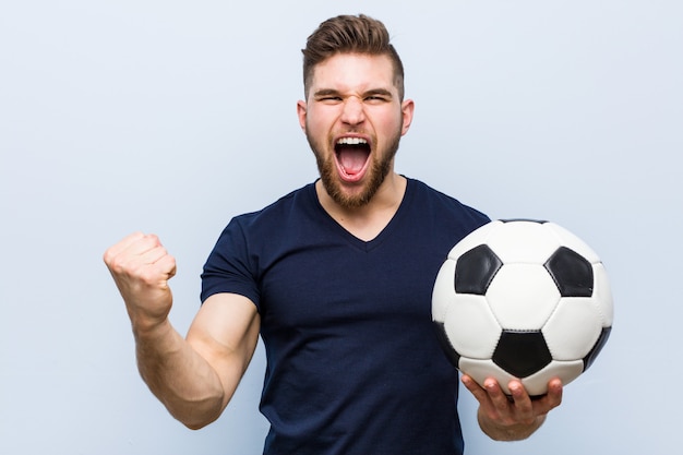 Jeune homme de race blanche tenant un ballon de football acclamant insouciant et excité.