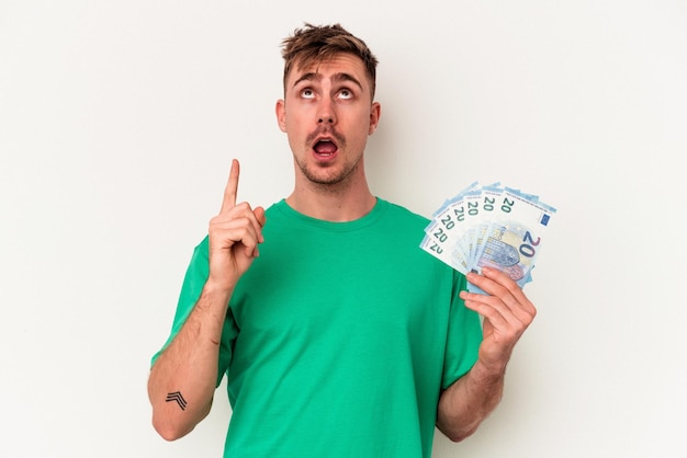 Jeune homme de race blanche tenant des billets de banque isolés sur fond blanc pointant vers le haut avec la bouche ouverte.