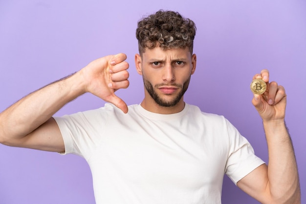 Jeune homme de race blanche tenant un Bitcoin isolé sur fond violet montrant le pouce vers le bas avec une expression négative