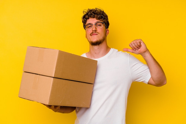 Jeune homme de race blanche tenant une boîte en carton isolée sur fond jaune se sent fier et confiant, exemple à suivre.