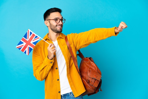 Jeune homme de race blanche tenant un drapeau du Royaume-Uni isolé sur fond jaune donnant un geste de pouce en l'air