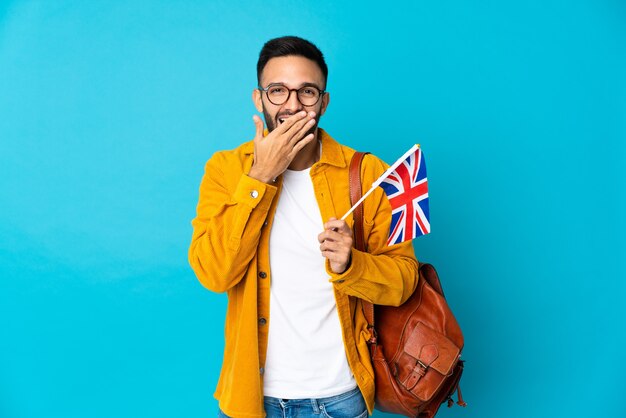 Jeune homme de race blanche tenant un drapeau du Royaume-Uni isolé sur fond jaune heureux et souriant couvrant la bouche avec la main