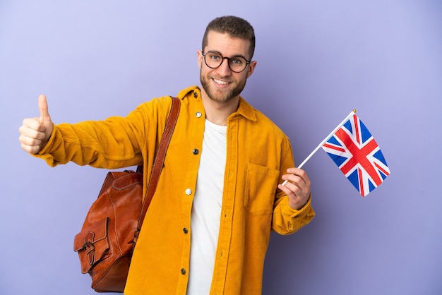 Jeune homme de race blanche tenant un drapeau du Royaume-Uni isolé sur un mur violet donnant un geste de pouce en l'air