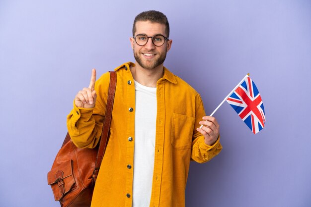 Jeune homme de race blanche tenant un drapeau du Royaume-Uni isolé sur un mur violet montrant et en soulevant un doigt