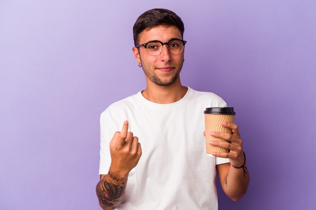 Jeune homme de race blanche tenant du café à emporter isolé sur fond violet pointant du doigt vers vous comme s'il vous invitait à vous rapprocher.