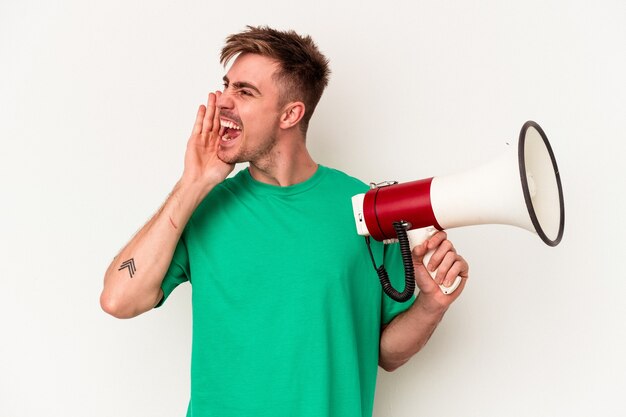 Jeune homme de race blanche tenant un mégaphone isolé sur fond blanc criant et tenant la paume près de la bouche ouverte.