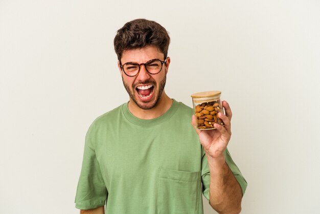 Jeune homme de race blanche tenant un pot d'amandes isolé sur fond blanc criant très en colère et agressif.