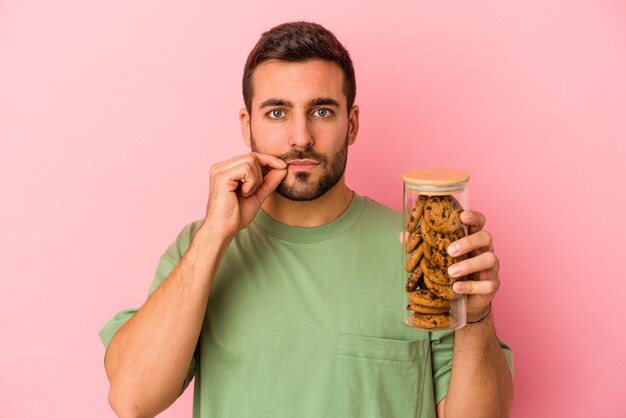 Jeune homme de race blanche tenant un pot de cookies isolé sur fond rose avec les doigts sur les lèvres gardant un secret.