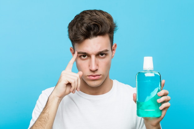 Jeune homme de race blanche tenant un rince-bouche pointant son temple avec le doigt, pensant, concentré sur une tâche.