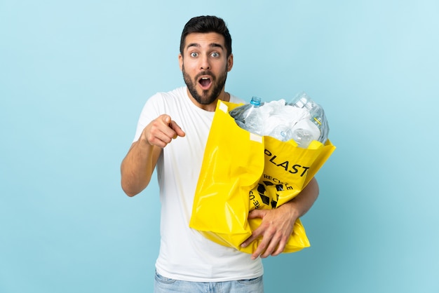 Jeune homme de race blanche tenant un sac plein de bouteilles en plastique à recycler isolé sur bleu surpris et pointant vers l'avant