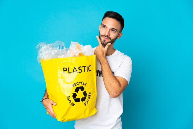 Jeune homme de race blanche tenant un sac plein de bouteilles en plastique à recycler isolé sur fond bleu ayant des doutes