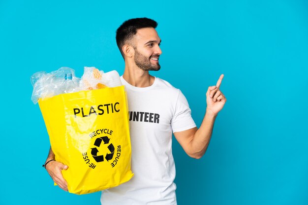 Jeune homme de race blanche tenant un sac plein de bouteilles en plastique à recycler isolé sur fond bleu pointant vers le haut une excellente idée