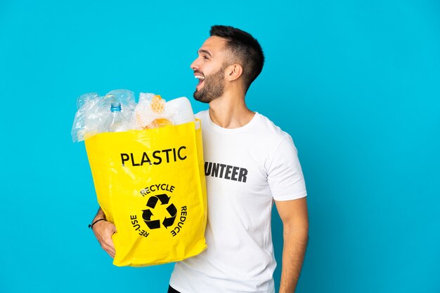 Jeune homme de race blanche tenant un sac plein de bouteilles en plastique à recycler isolé sur fond bleu en riant en position latérale