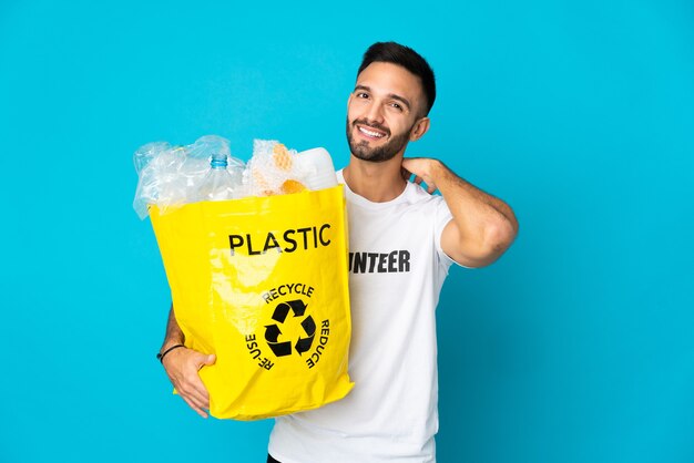 Jeune homme de race blanche tenant un sac plein de bouteilles en plastique à recycler isolé sur fond bleu en riant