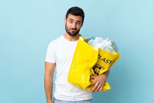 Jeune homme de race blanche tenant un sac plein de bouteilles en plastique à recycler isolé sur mur bleu avec une expression triste