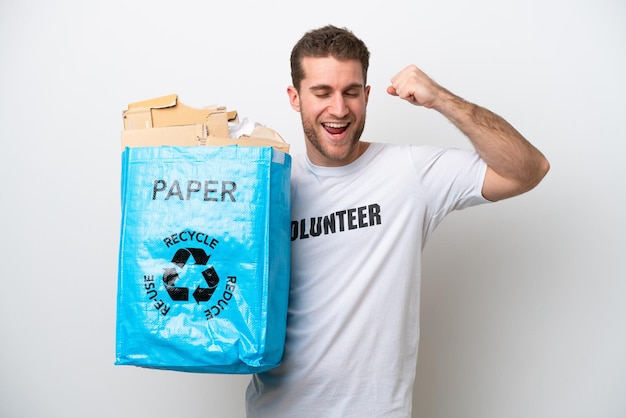 Jeune homme de race blanche tenant un sac de recyclage plein de papier à recycler isolé sur fond blanc célébrant une victoire