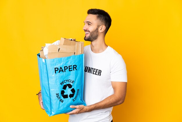 Jeune homme de race blanche tenant un sac de recyclage plein de papier à recycler isolé sur fond blanc à côté