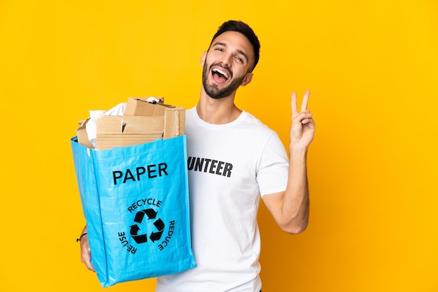 Jeune Homme De Race Blanche Tenant Un Sac De Recyclage Plein De Papier à Recycler Isolé Sur Fond Blanc Souriant Et Montrant Le Signe De La Victoire