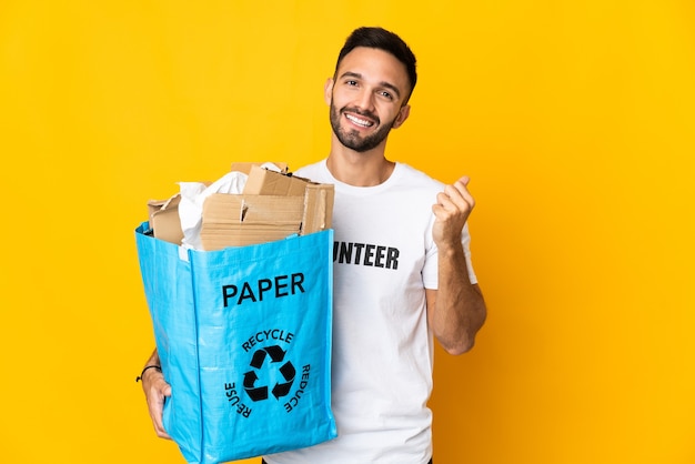 Jeune homme de race blanche tenant un sac de recyclage plein de papier à recycler isolé sur un mur blanc faisant le geste de l'argent