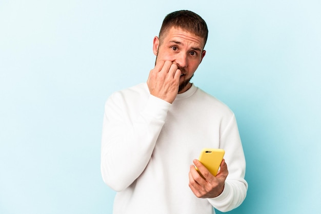 Jeune homme de race blanche tenant un téléphone portable isolé sur fond bleu se rongeant les ongles, nerveux et très anxieux.