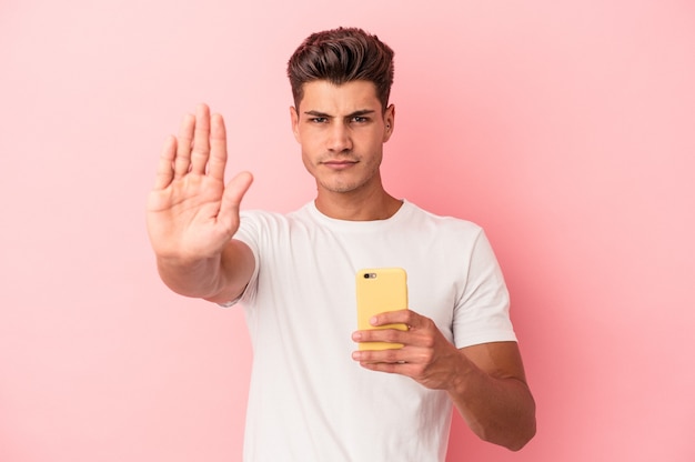 Jeune homme de race blanche tenant un téléphone portable isolé sur fond rose debout avec la main tendue montrant un panneau d'arrêt, vous empêchant.