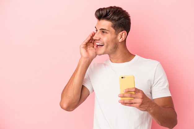 Un jeune homme de race blanche tenant un téléphone portable isolé sur fond rose dit une nouvelle secrète de freinage à chaud et regarde de côté