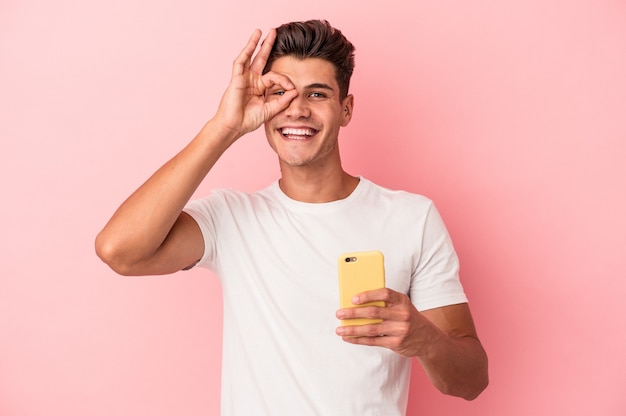 Jeune homme de race blanche tenant un téléphone portable isolé sur fond rose excité en gardant un geste ok sur les yeux.