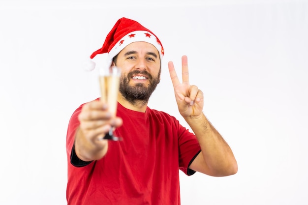 Jeune homme de race blanche très heureux avec un chapeau de Noël rouge grillant avec un verre de champagne sur fond blanc, vêtu d'une chemise rouge