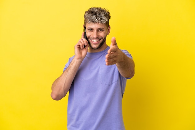 Jeune homme de race blanche utilisant un téléphone portable isolé sur fond jaune se serrant la main pour conclure une bonne affaire