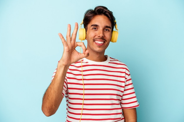 Jeune homme de race mixte écoutant de la musique isolée sur fond bleu joyeux et confiant montrant un geste correct.