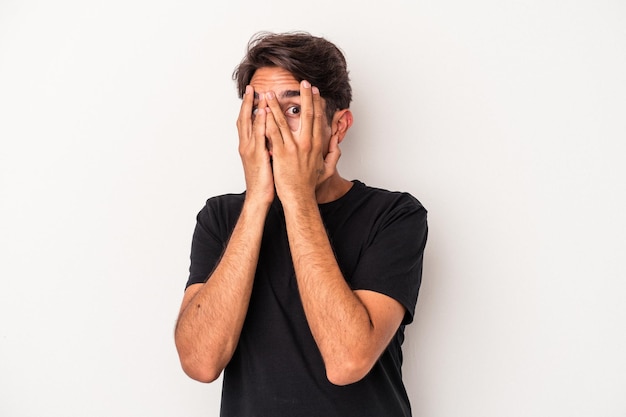 Jeune homme de race mixte isolé sur fond blanc clignote à travers les doigts effrayé et nerveux.