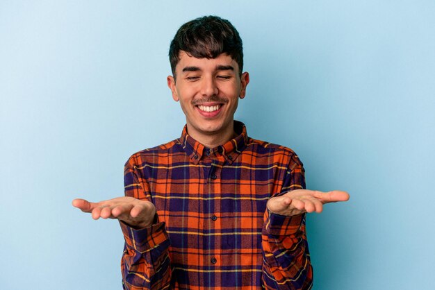 Jeune homme de race mixte isolé sur fond bleu tenant quelque chose avec des paumes, offrant à la caméra.