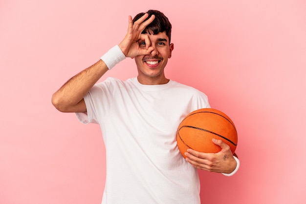 Jeune homme de race mixte jouant au basket-ball isolé sur fond rose excité en gardant le geste ok sur les yeux.