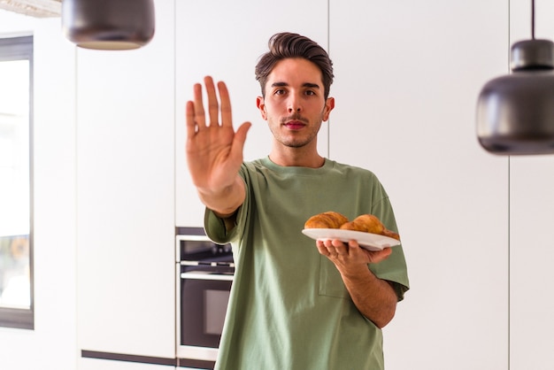 Jeune homme de race mixte mangeant un croissant dans une cuisine le matin debout avec la main tendue montrant un panneau d'arrêt, vous empêchant.