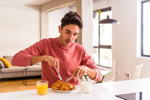 Jeune homme de race mixte mangeant un croissant dans une cuisine le matin