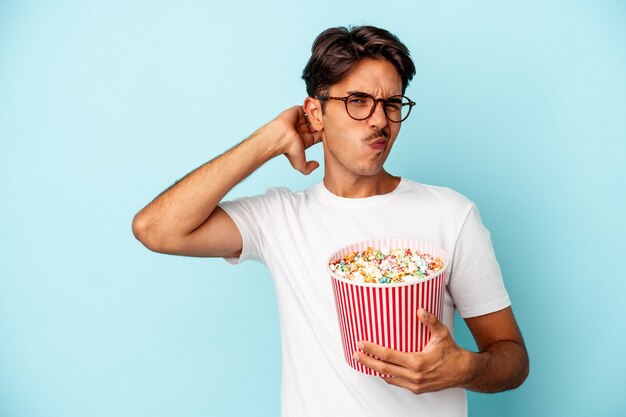 Jeune homme de race mixte mangeant des pop-corns isolés sur fond bleu touchant l'arrière de la tête, pensant et faisant un choix.