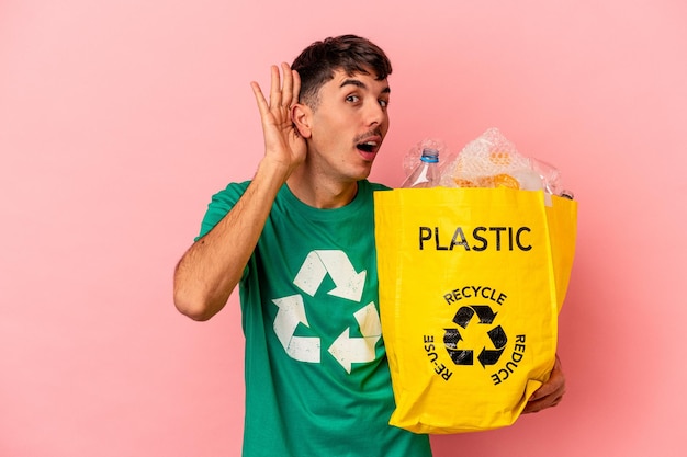 Jeune homme de race mixte en plastique recyclé isolé sur fond rose essayant d'écouter un potin.