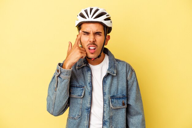 Jeune homme de race mixte portant un casque vélo isolé sur fond jaune montrant un geste de déception avec l'index.