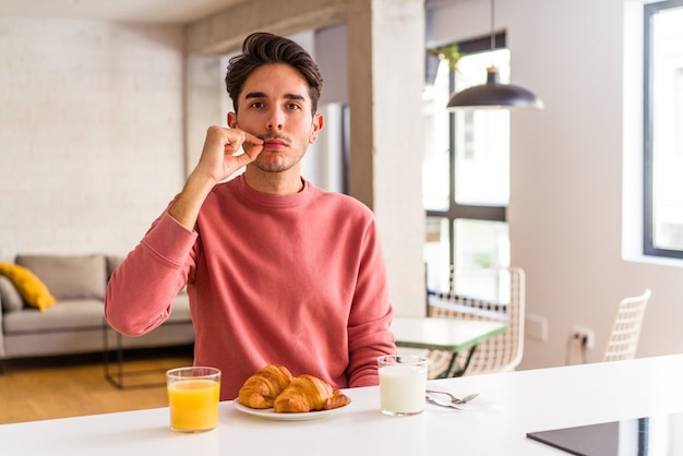 Jeune homme de race mixte prenant son petit déjeuner dans une cuisine le matin avec les doigts sur les lèvres gardant un secret.