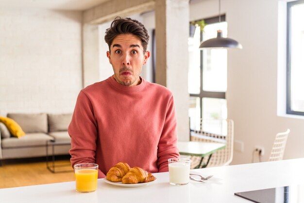 Un jeune homme de race mixte prenant son petit-déjeuner dans une cuisine le matin hausse les épaules et ouvre les yeux confus.