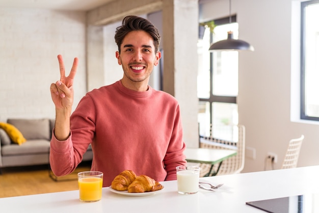 Jeune homme de race mixte prenant son petit déjeuner dans une cuisine le matin montrant le numéro deux avec les doigts.