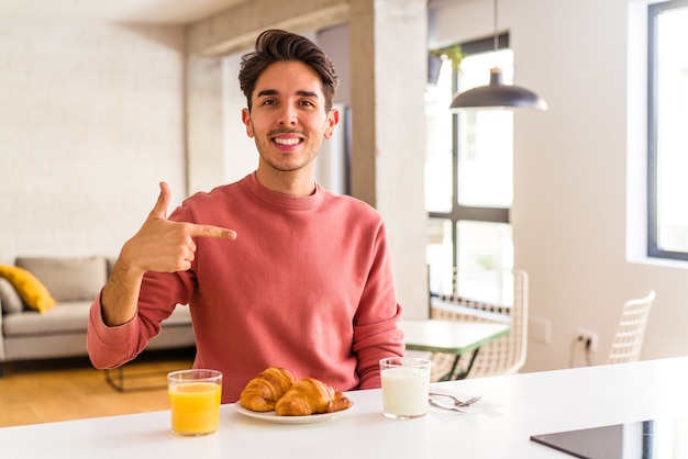 Jeune homme de race mixte prenant son petit déjeuner dans une cuisine le matin personne pointant à la main vers un espace de copie de chemise, fier et confiant