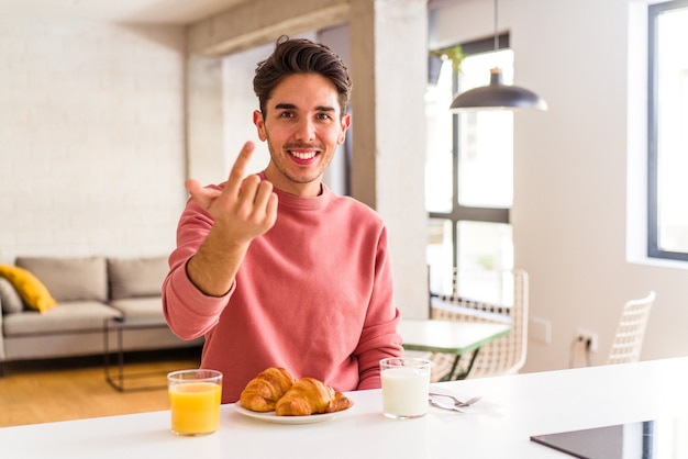 Jeune homme de race mixte prenant son petit-déjeuner dans une cuisine le matin en vous pointant du doigt comme s'il vous invitait à vous rapprocher.