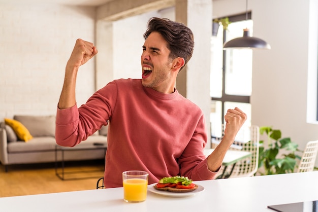 Jeune homme de race mixte prenant son petit déjeuner dans sa cuisine levant le poing après une victoire, concept gagnant.