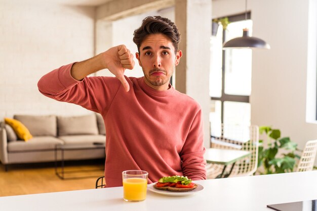 Jeune homme de race mixte prenant son petit déjeuner dans sa cuisine montrant un geste d'aversion, les pouces vers le bas. Notion de désaccord.