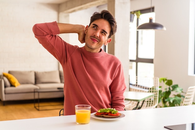 Jeune homme de race mixte prenant son petit déjeuner dans sa cuisine touchant l'arrière de la tête, pensant et faisant un choix.