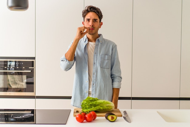 Jeune homme de race mixte préparant une salade pour le déjeuner avec les doigts sur les lèvres en gardant un secret.