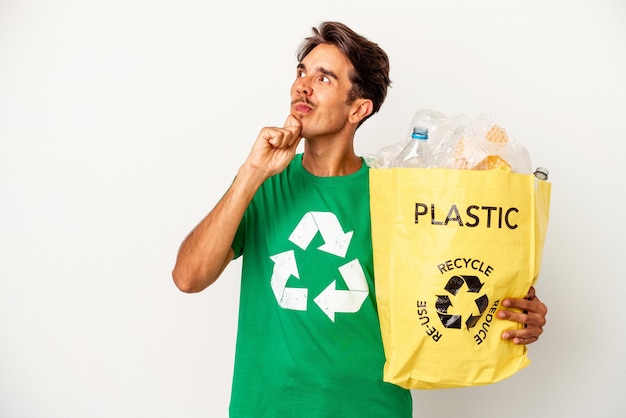 Jeune homme de race mixte recyclant du plastique isolé sur fond jaune regardant de côté avec une expression douteuse et sceptique.